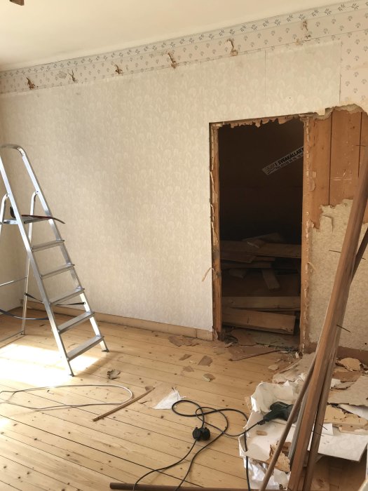 Renoveringsarbete i rum med trägolv, tapetserade väggar och rivet tak med synlig isolering. Stege och rivningsavfall syns.
