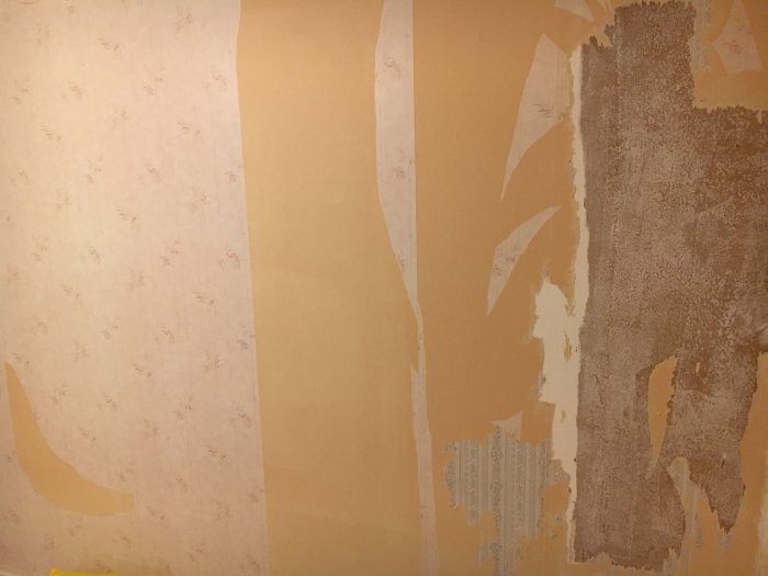 Vägg med delvis bortriven tapet i olika lager, synligt spackle-arbete och ojämnheter.