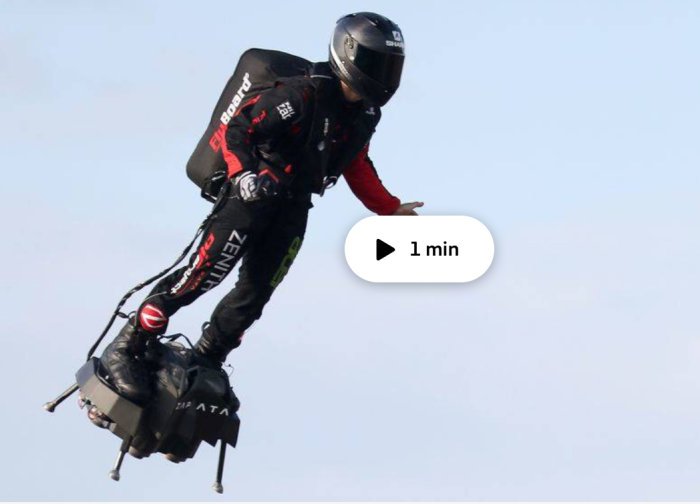 Person i svart röd klädsel och hjälm flyger med en ryggburen jetpack mot en klar himmel.