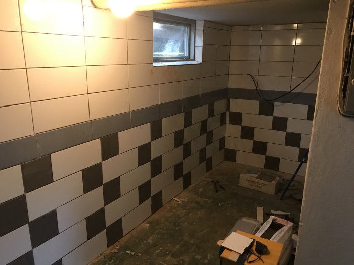 Orenoverad källare under förvandling med delvis lagd svartvit klinker på väggarna, byggmaterial på golvet och ett litet fönster.