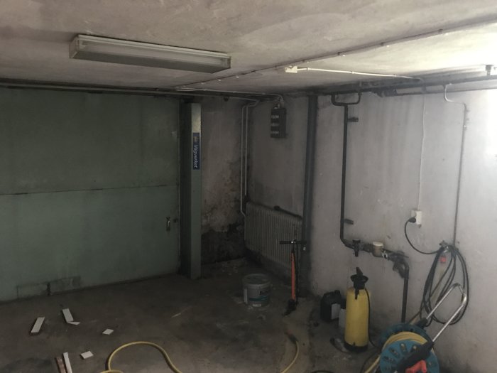 Orenoverad källare med betongväggar, rörledningar och en garageport.