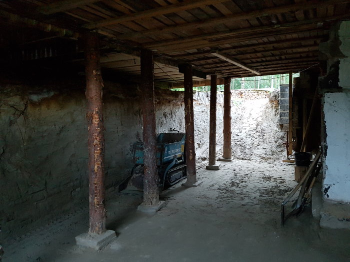 Underarbete under ett hus med stödbjälkar och en självlastande grävare, visar grunden under ombyggnad.