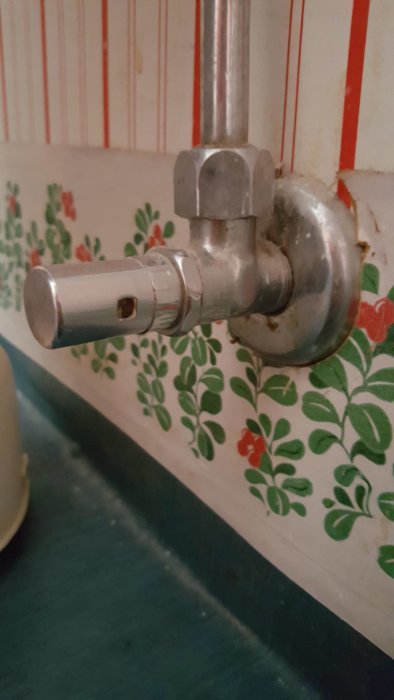 Gammal avstängningsventil för vatten, silverfärgad, mot mönstrad tapet.