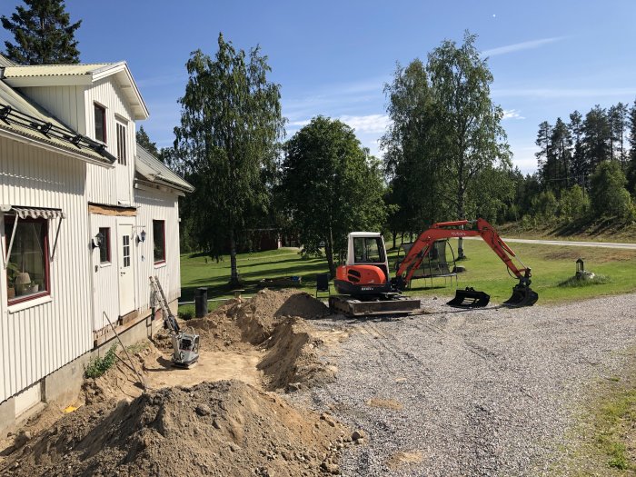 Grävmaskin och grushög vid vit husgrund på solig dag, grävarbete i trädgård.