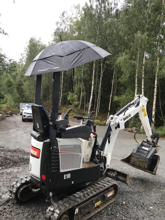 En minigrävare Bobcat E10 med ett stort paraply fäst ovanpå i en skogsmiljö.