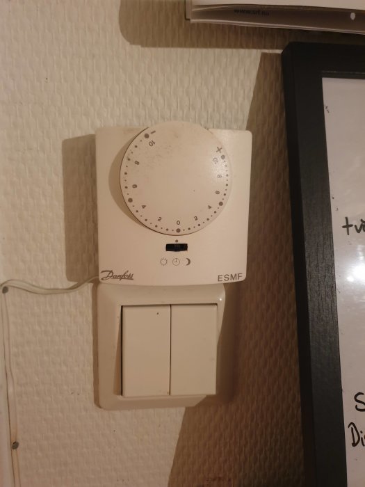 Väggmonterad Danfoss termostat med skruvreglage ovanför två ljusströmbrytare, använd för värmeinställningar.