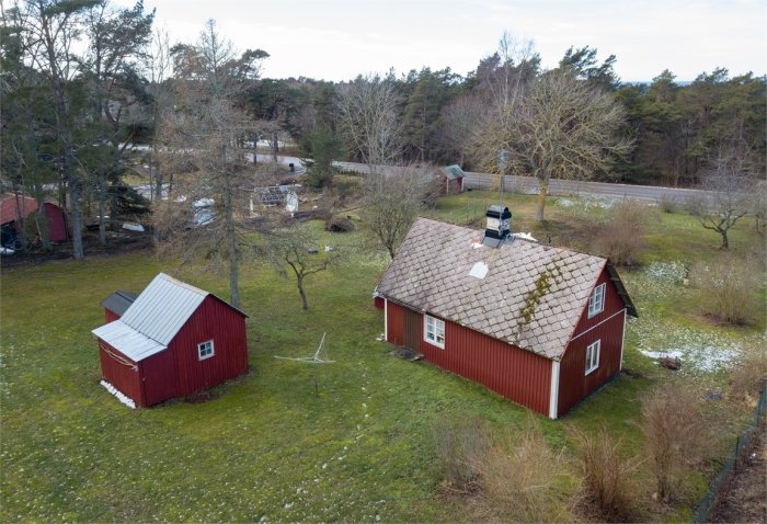 Flygbild av ett rödmålat torp med tegeltak och omgivande grönt landskap, marker som sluttar mot huset.