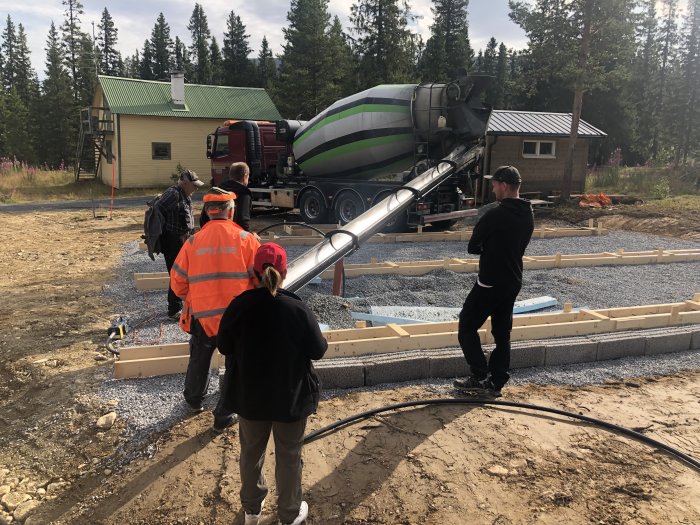 Betongbil häller betong i gjutformar för husgrunder med arbetare på en byggplats med skogsbakgrund.