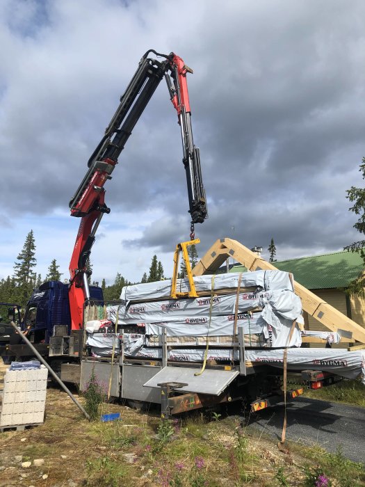 Lastbil med kran lyfter byggmaterial vid en konstruktion med inslag av lecablock och trästomme under uppbyggnad.