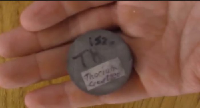 Hand som håller en rund grå metallbit märkt med "232 Th Thorium Ceramic".