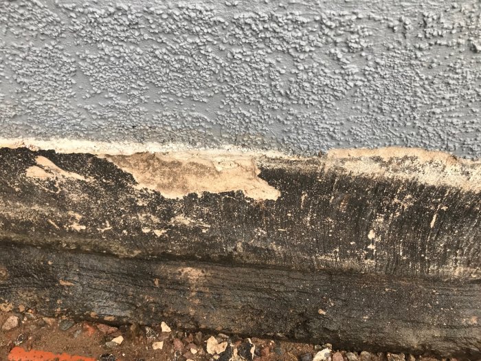 Närbild av skadad grundmur med avflagande kallasfalt och synlig betong och sten.