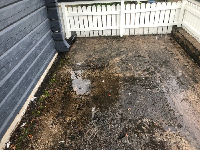 Våt mark och vattenpölar bredvid trästaket och grått hus efter regn.