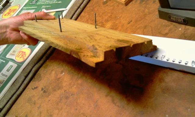 Et stykke trykimprægneret træ med søm og udskårne kanter, placeret på et bord ved siden af en målepind, holdt af en hånd.