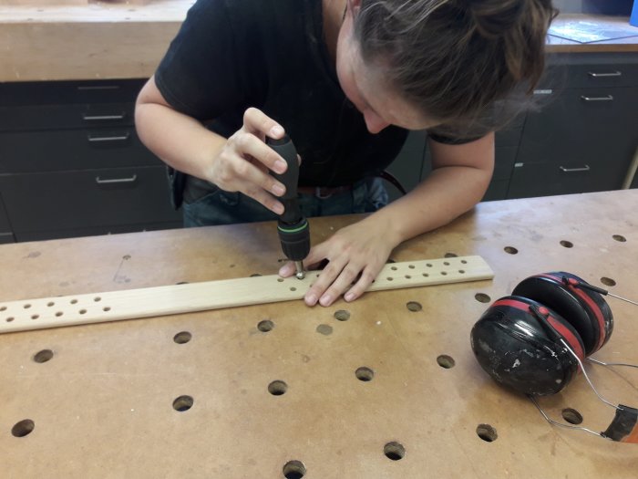 Person som arbetar på en träspjäla med borr och försänkare, hörskydd ligger på arbetsbordet.