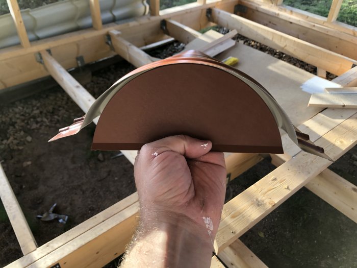Hand håller ett brunt ändlock för nockplåt framför ett pågående bygge av uthus.