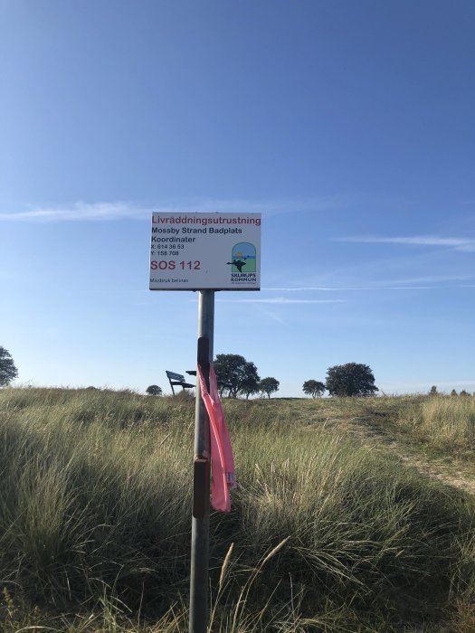 Skylt om livräddningsutrustning med en rosa baddräkt hängande på stolpen, naturlig bakgrund.