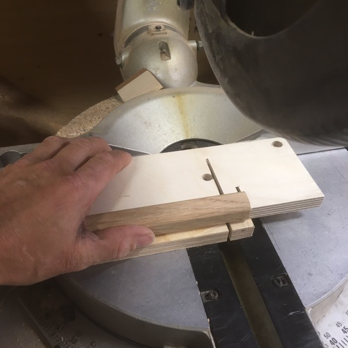 Hand holding a small wooden block against a jig on a miter saw table, preparing to make a precise cut.