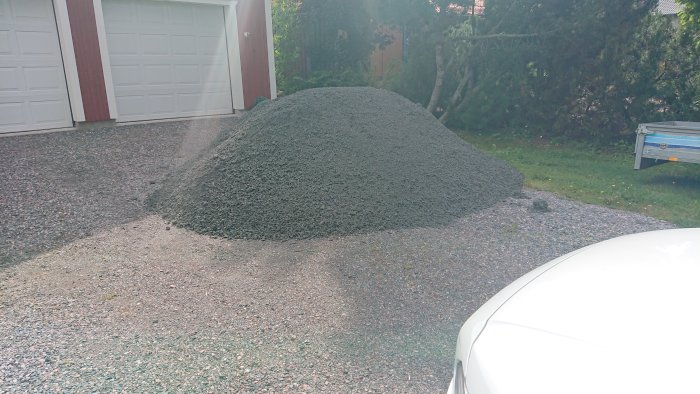 Stor hög med grus framför garage på uppfart, delvis täckt av skugga, med släpvagn i bakgrunden.