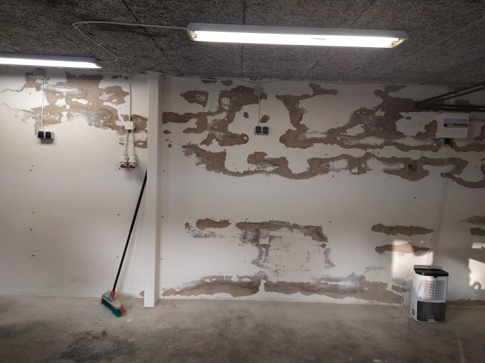 Garagevägg med flagnad puts på Lecablock, synliga reparationer och avfuktare på golvet.