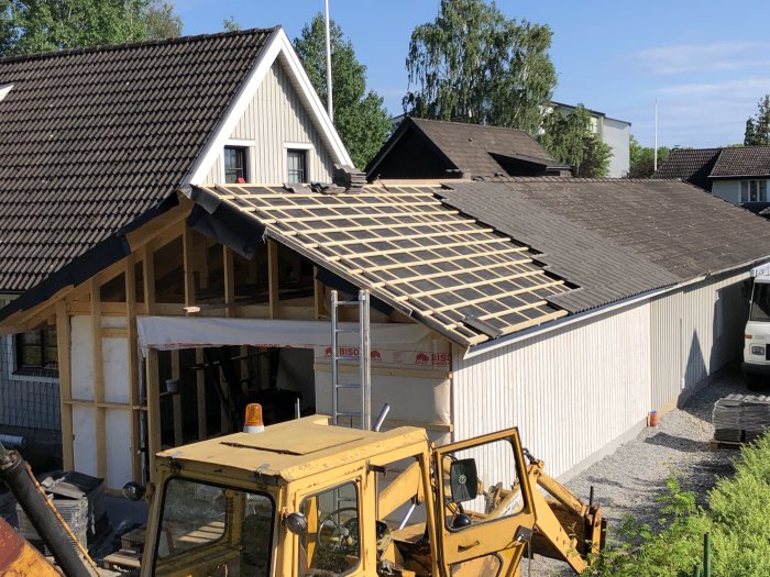 Pågående takrenovering på en villa med ett delvis lagt träregelverk och en gul grävmaskin i förgrunden.