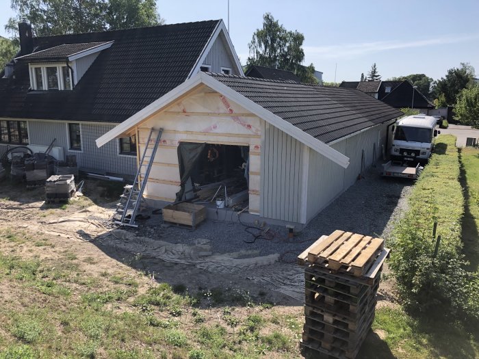 Nybyggnation av en fristående garagebyggnad med öppen port och synliga träreglar, nära ett bostadshus.