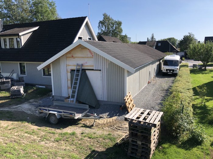 Renoveringsarbete på en husfasad med en stege, byggmaterial och släpvagn framför huset.