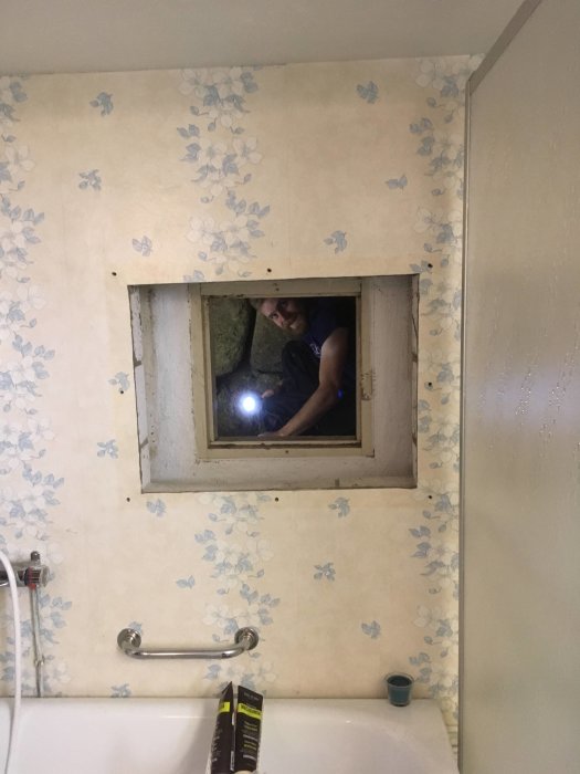 Person tittar genom lucka i vägg från krypgrund in till en tvättstuga med mönstrad tapet.