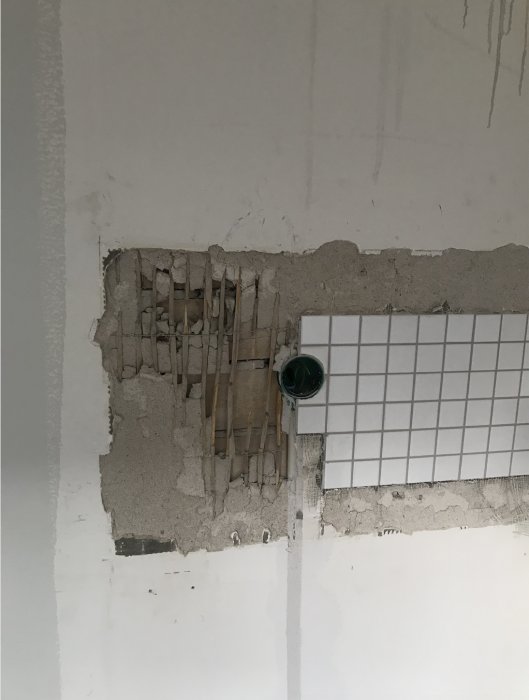 Skadad vägg där gammalt kakel tagits bort, visande oskyddad murbruk och en grön elrörskoppling.