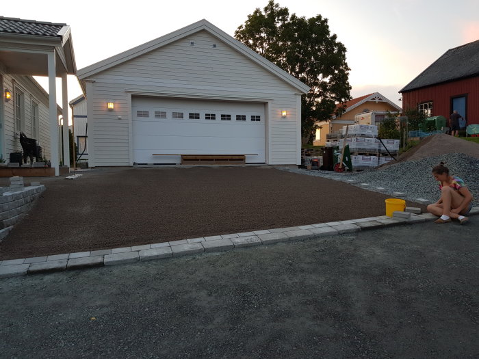 Nyjämnad grusyta framför vit garagebyggnad i förberedelse för stensättning med person sittandes vid sidan.