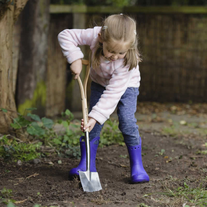 Flicka med rosa tröja och blå stövlar gräver i jord med en spade.