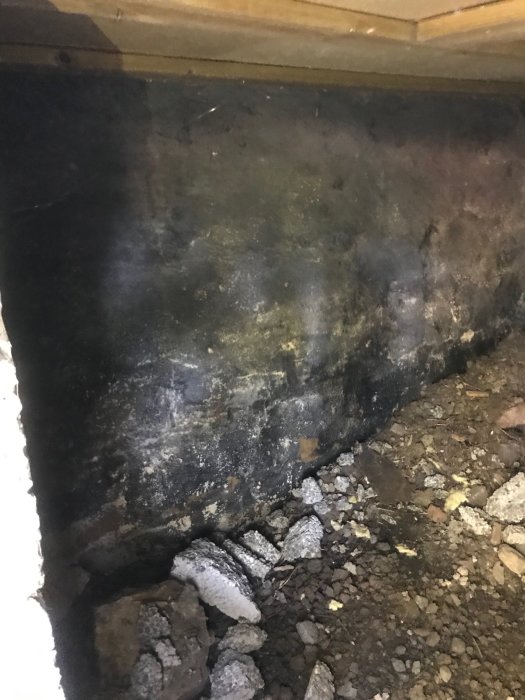 Fuktig krypgrund med synliga fuktfläckar och kondens på svarta plastmattan, grus och stenar på marken.