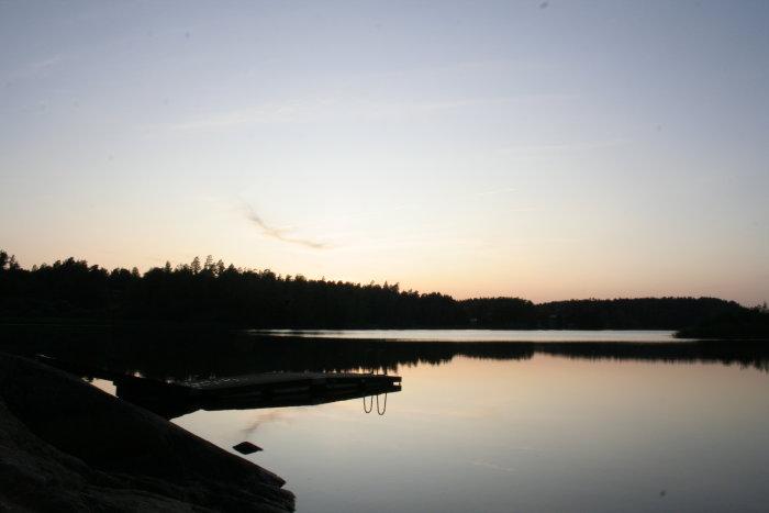 Skymning över en stilla Långsjö med reflekterande vatten och en brygga mot siluett av skog.