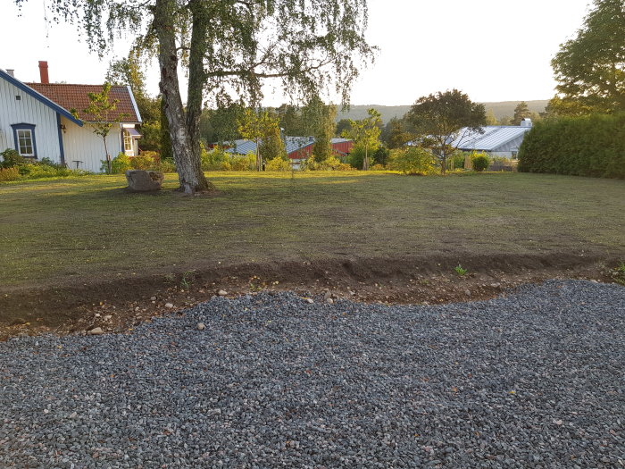 Trädgård under groning med sparsamt gräs och stor sten framför huset, nyanlagt grus i förgrunden.