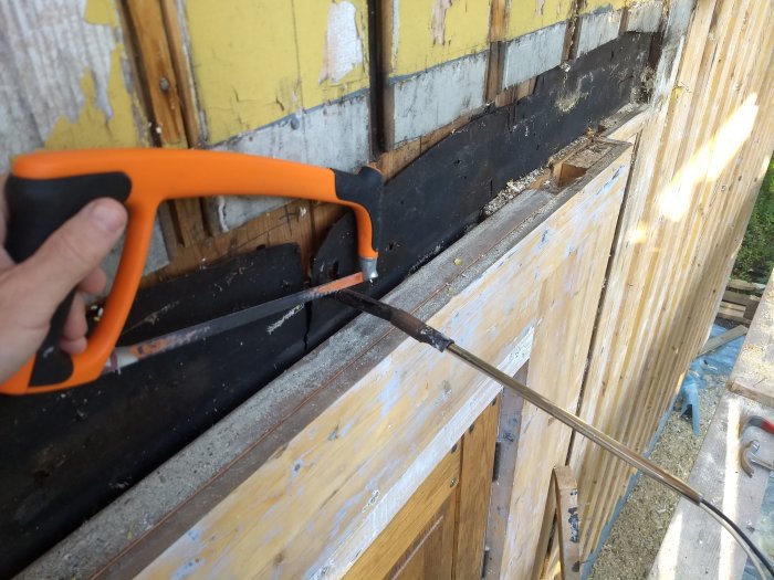 Person som sågar genom en träplanka med en handsåg vid en husvägg under renovering.