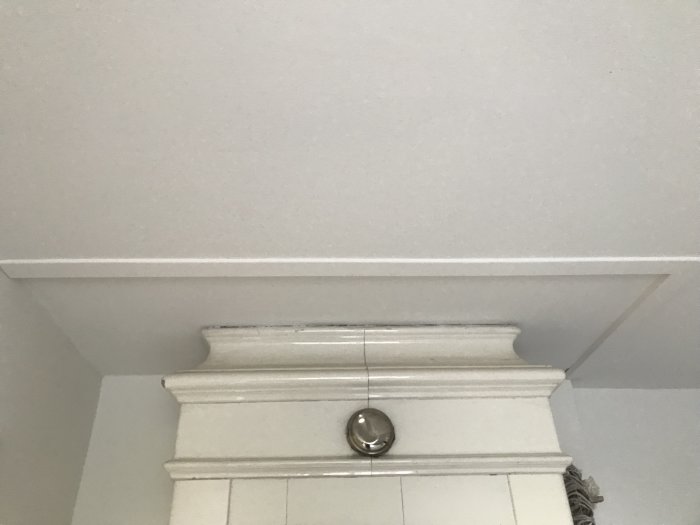 Vit kakelugn med ornamentik och en liten springa till det vitmålade taket ovanför.