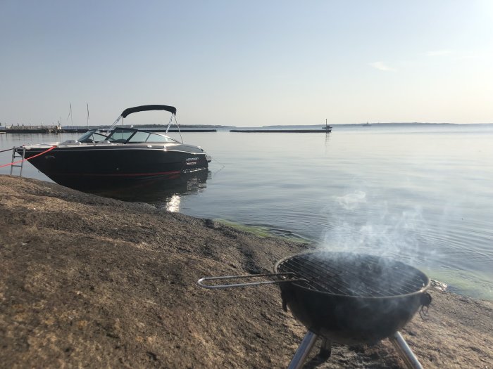 Båt förtöjd vid klippa med grill i förgrunden och lugnt hav i solnedgång.