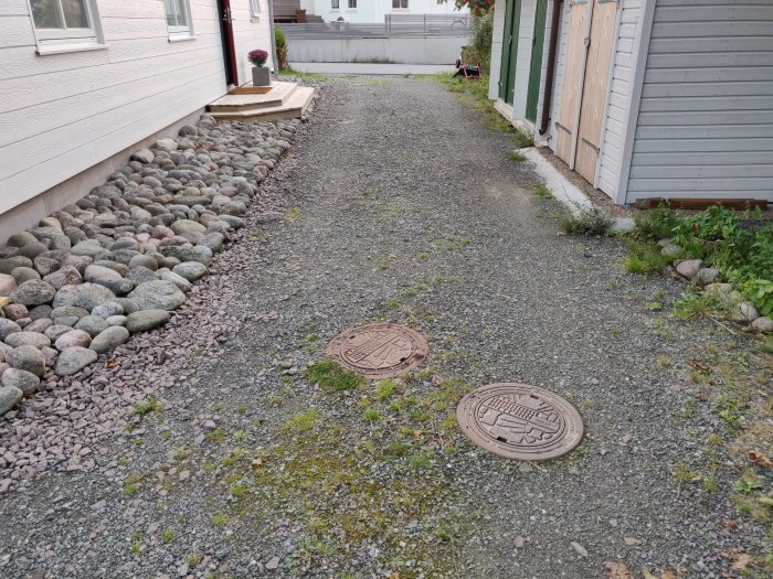 Grusväg med ogräs utanför hus med kullerstenskant och två brunnslock.