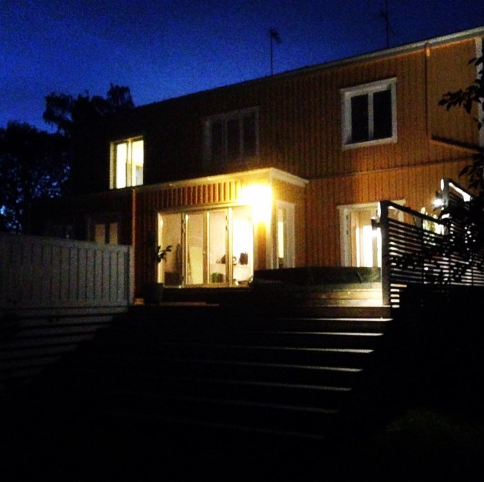 Ett tvåvåningshus med tända fönster och balkongdörr på kvällen.
