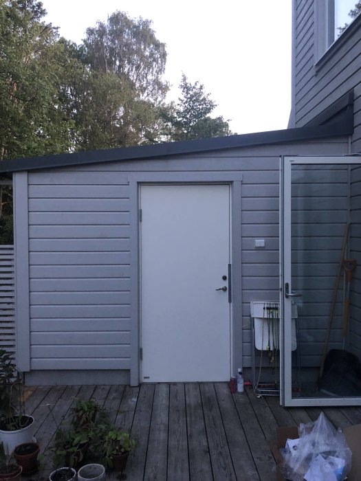 En liten grå skjul bredvid ett hus, dörr stängd, omringad av krukväxter och trädäck, vid skymning.