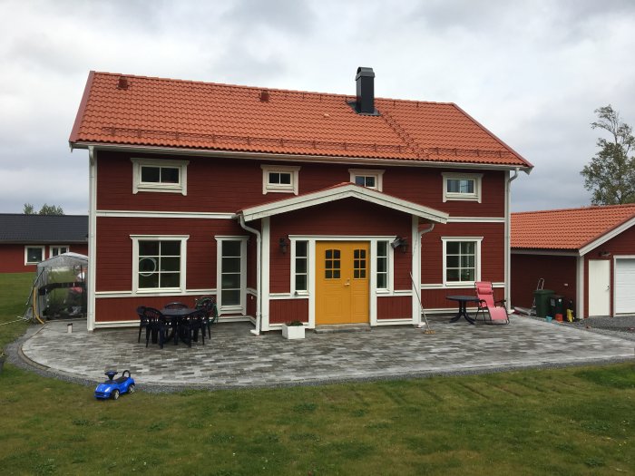 Ny stenläggning framför ett rött hus med gult dörr och tegeltak, möbler och leksaker på gräsmattan.