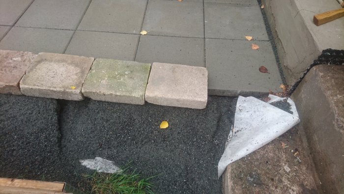 Upphöjd kant av trottoarstenar vid en uteplats, där ett underlag av stenmjöl syns, tydande på erosion.