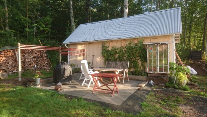 Ett uteområde med ett litet träskjul, möblerad terrass och vedstapel vid skogsbryn i solljus.
