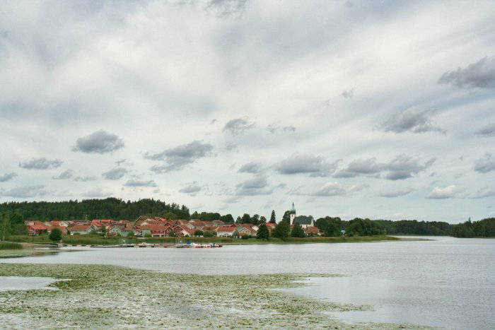 Utsikt över Frösjön med Frustuna kyrka i bakgrunden och röda hus i förgrunden, molnig himmel ovan.