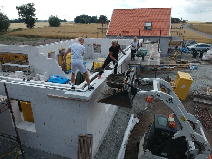 Personer jobbar på en byggarbetsplats, lägger tegel och använder en grävmaskin, med pågående husbygge i bakgrund.