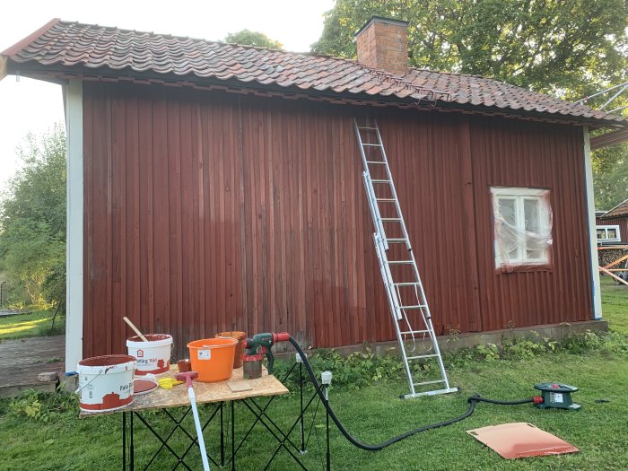 Nytt målarprojekt visas med en stege lutad mot en nymålad röd trävägg, målarutrustning och hinkar på marken.