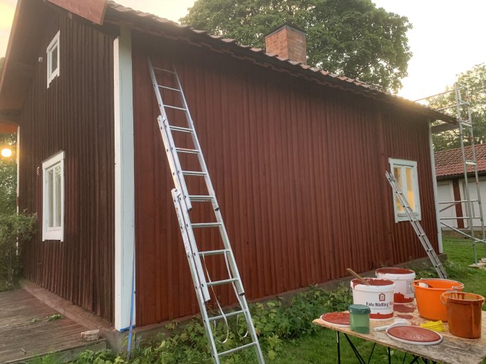 Nyfärgad röd träfasad på hus med stege och målarutrustning framför i skymningen.