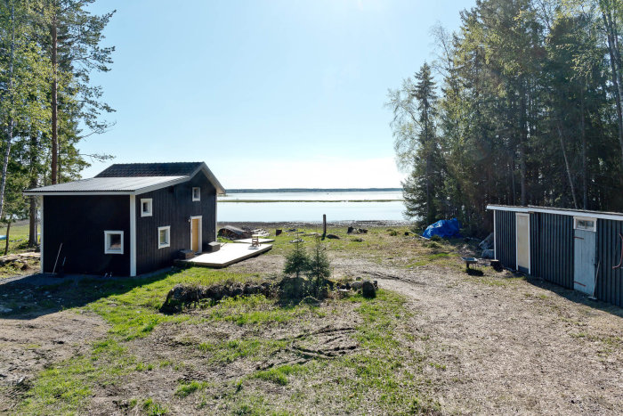 Tomt vid vattnet med ojämn gräsmatta, stenbumlingar och ett svart hus med blandat tak.