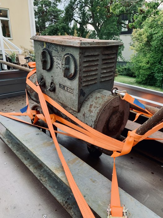 En gammal Esab K200 omformare uppbunden med orange spännband på en släpvagn bredvid i-balkar och en lång stång.