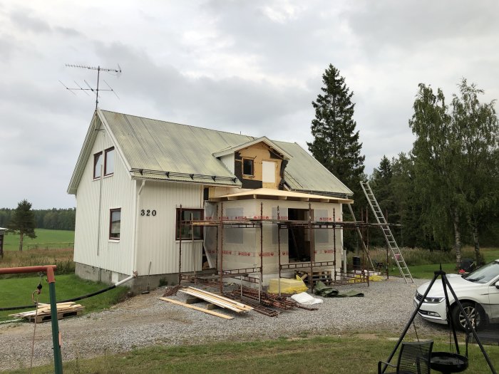 Hus under renovering med ny farstukvist i stomme och valmat tak under arbete, omgiven av träd och gräsmatta.