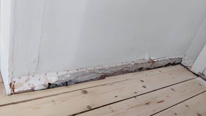 Skadad vägg och borttagen list där putsen fallit bort längs med trägolvet.